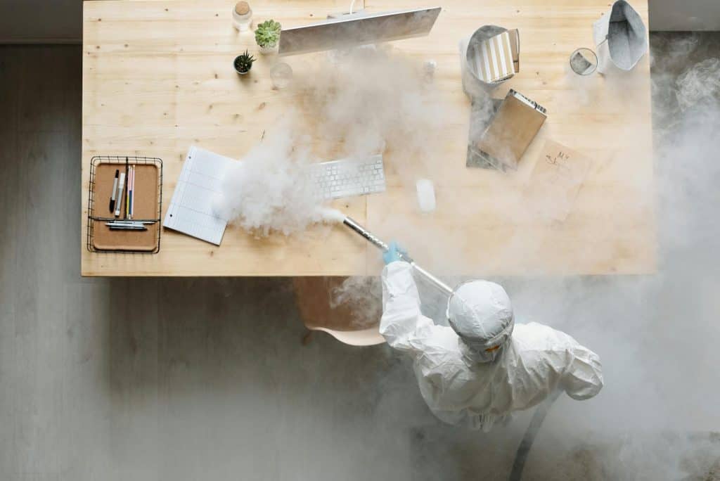 Photo by Michelangelo Buonarroti An individual disinfects a workspace using fumigation, wearing full protective gear for safety.